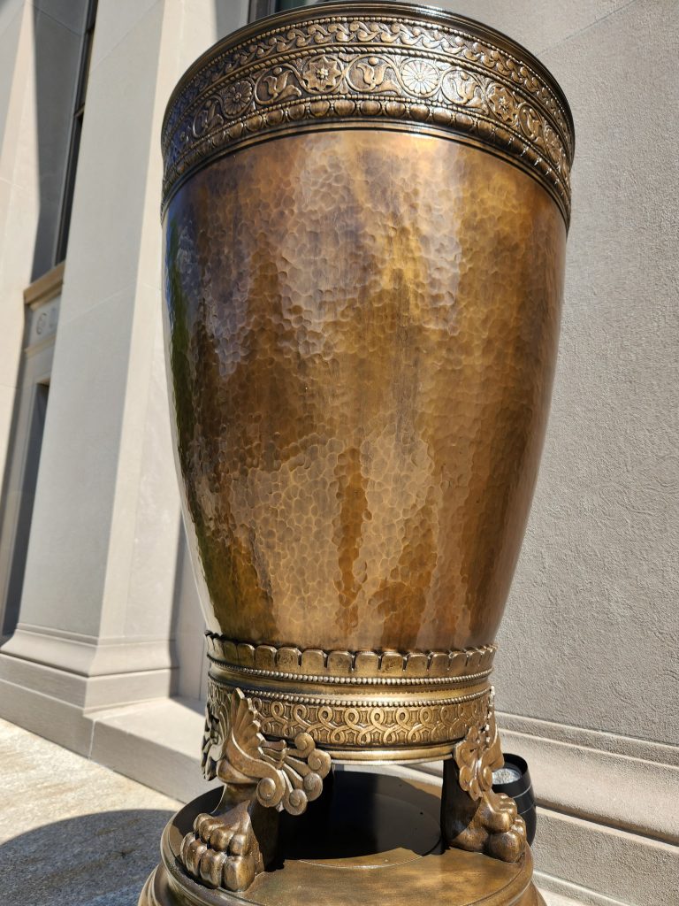 Close-up of restored hammered bronze urn with ornate trim and pedestal base outside a civic building